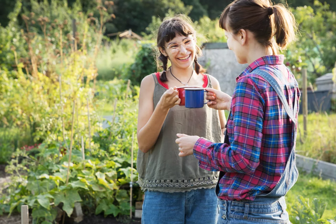 Two young women, both wearing casual clothes, smile and clink colorful mugs together in a sunny garden setting. The scene evokes friendship, relaxation, and shared moments in nature.
