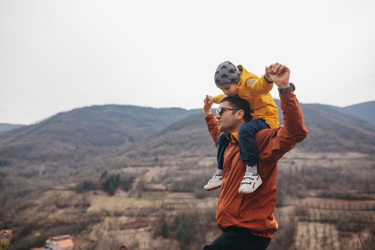 Young father carrying his little son in yellow jacket and cap on shoulders,standing together and enjoying the view from top of the mountain