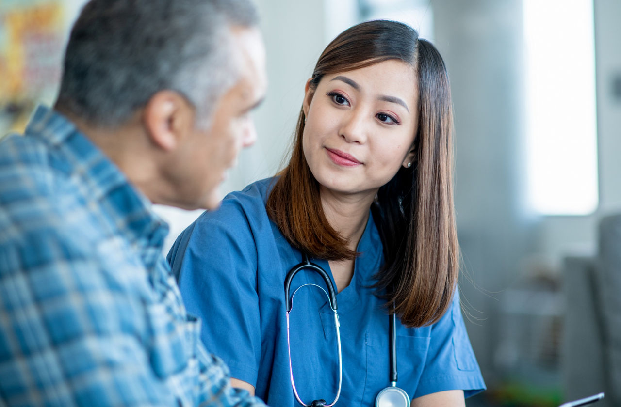 A female nurse of Asian decent makes a home care visit to a senior patient.  She is wearing blue scrubs and holding out a tablet as she reviews recent test results with the senior.