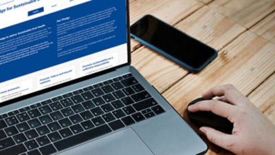 Corner of a keyboard and laptop screen showing sustainability pledge, plus a hand on a mouse and a mobile phone on the right