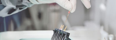Gloved hand holding rotary endodontic files above a sterilization or preparation tray in a dental clinic setting.