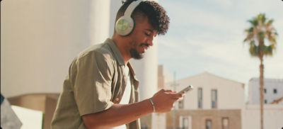 A man wearing headphones looks at his phone while standing on a balcony overlooking a sunny city.