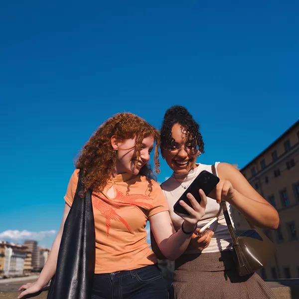 Two friends laughing together while looking at a smartphone on a sunny day in the city. 