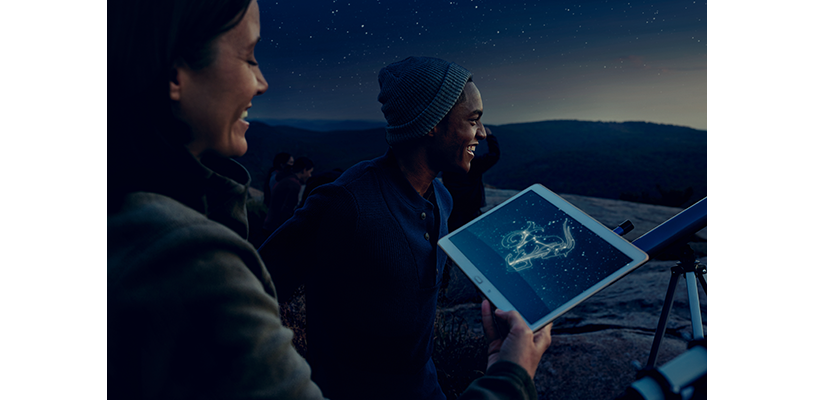 Two astronomers look at night sky and hold a mobile tablet