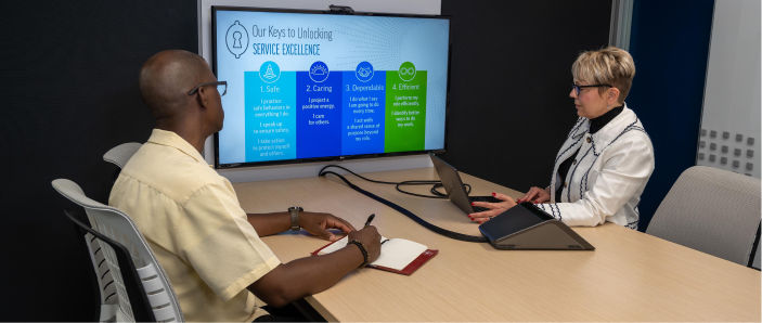 A man and woman sitting at a table looking at a work presentation on a television screen.