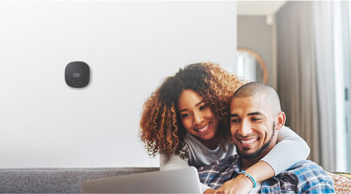 Couple smiling and looking at computer with a smart thermostat in the background