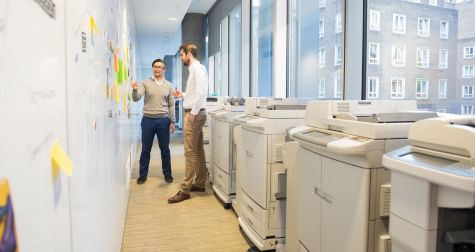 Two IT people, discussing work, in a halfway filled with printers.