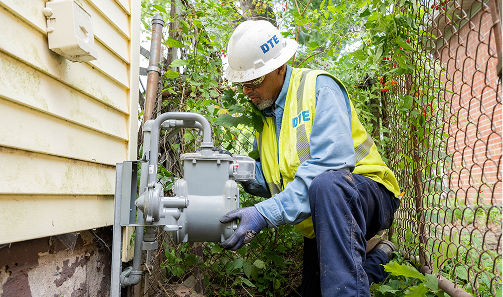 DTE employee with hard hat, yellow vest and gloves, inspecting a natural gas meter outdoors.