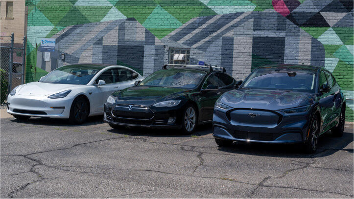 Three electric cars parked in a business lot, with a vibrant mural decorating the wall behind them.