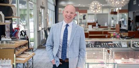 Man wearing a blue suit and tie smiling at camera standing inside a bright jewelry store.