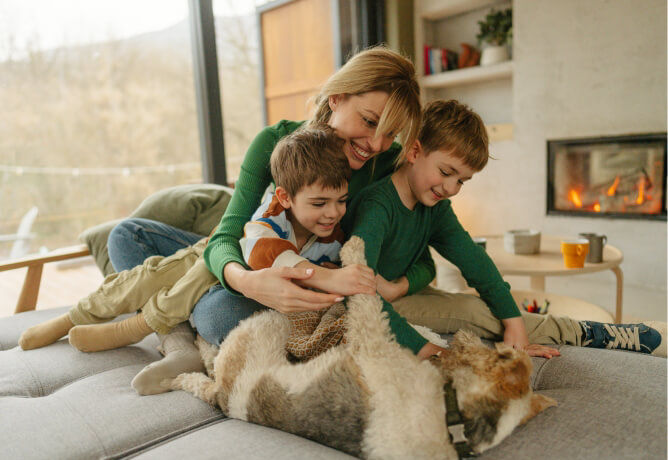 Mom with two kids playing with a dog in a living room with fireplace in the background.