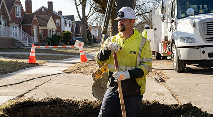 DTE employee with safety gear holding a shovel at a residential work site with traffic cones, barriers, and a white utility truck in the background.