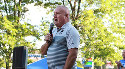 Older man speaking at an outdoor event with green trees in the background.