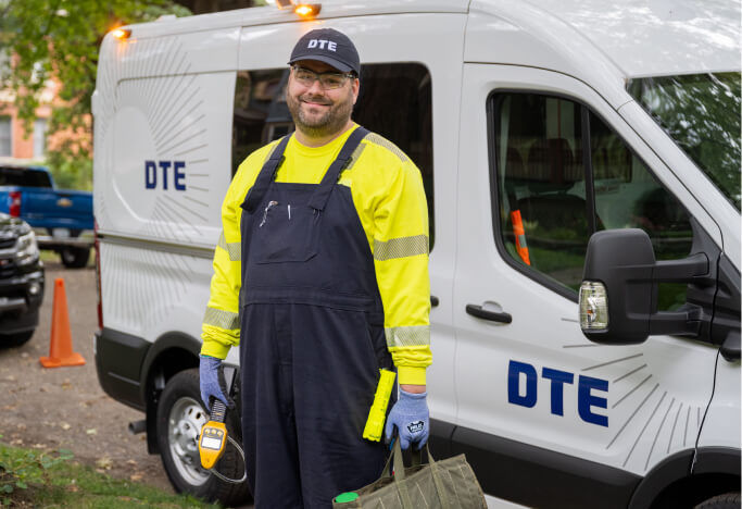 DTE employee wearing uniform and smiling at camera with a DTE while van behind him.