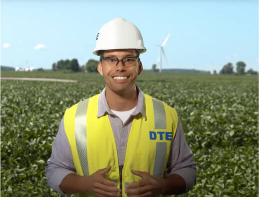 DTE employee wearing a hard hat and smiling standing in front of a corn field and wind turbine