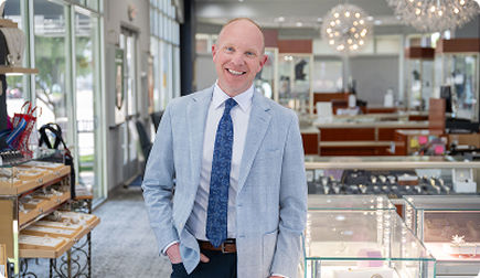 Man wearing light blue suit smiling at camera inside a jewelry store.