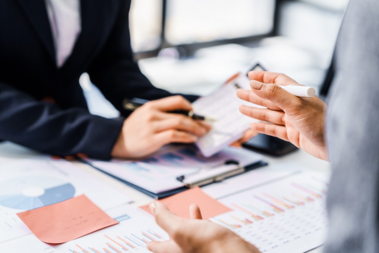 Two female business professionals in suits sit at desk, engaged in strategic discussion. They analyze financial planning, risk management, corporate growth while reviewing reports and market trends.