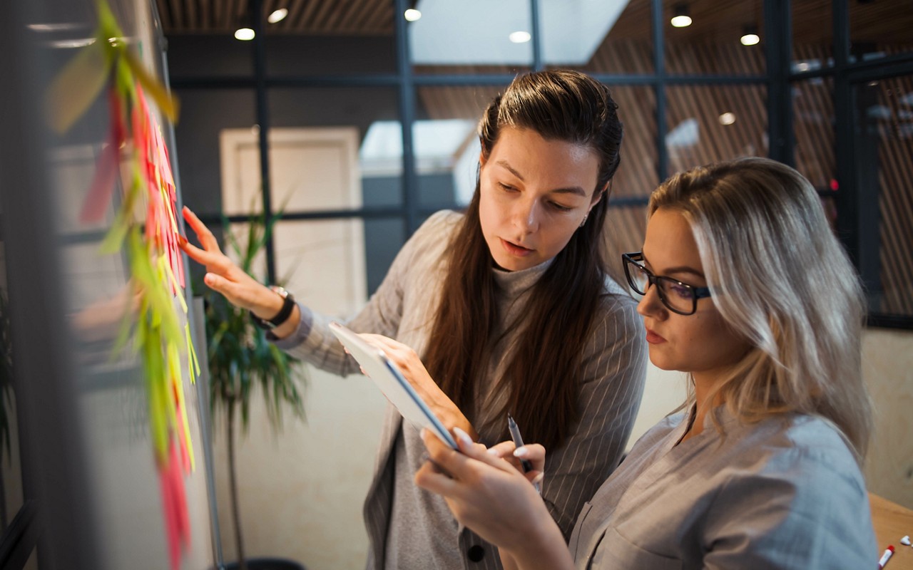 Teamwork, project management, agile methodology. Two young business women in the office are planning product development and support. Colleagues glue sticky stickers on the Board