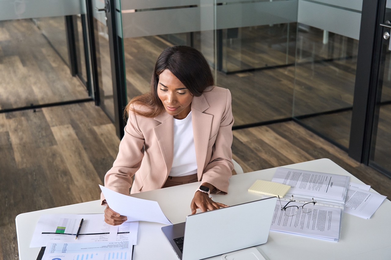 Busy African American business woman professional accountant, marketing manager, financial executive checking document report using laptop checking company papers working sitting at office desk.