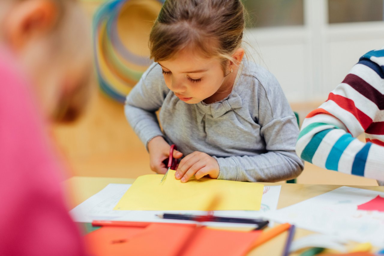 A young child carefully cutting a piece of yellow construction paper with safety scissors at a table.