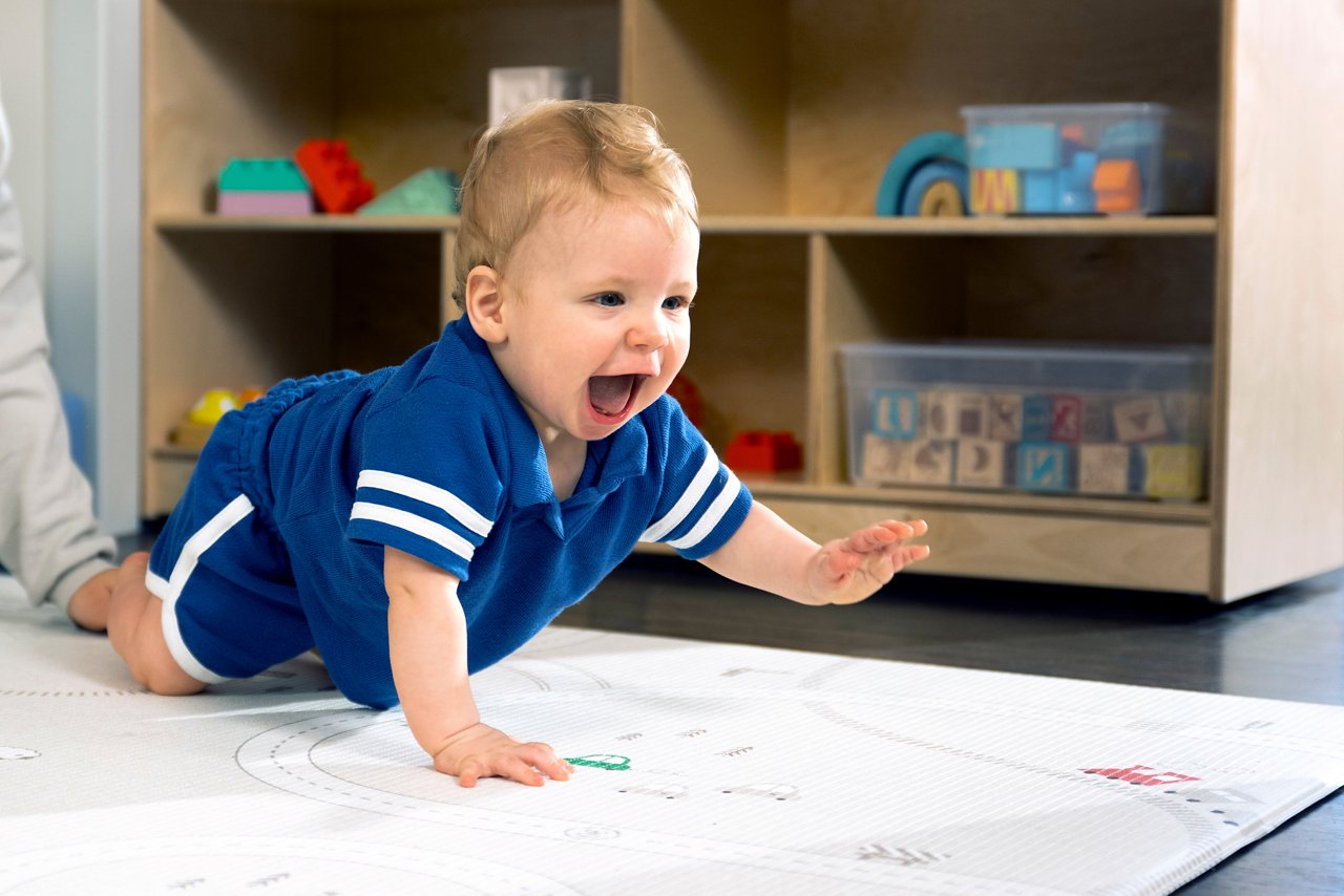 A smiling baby crawling across the floor in a preschool classroom.