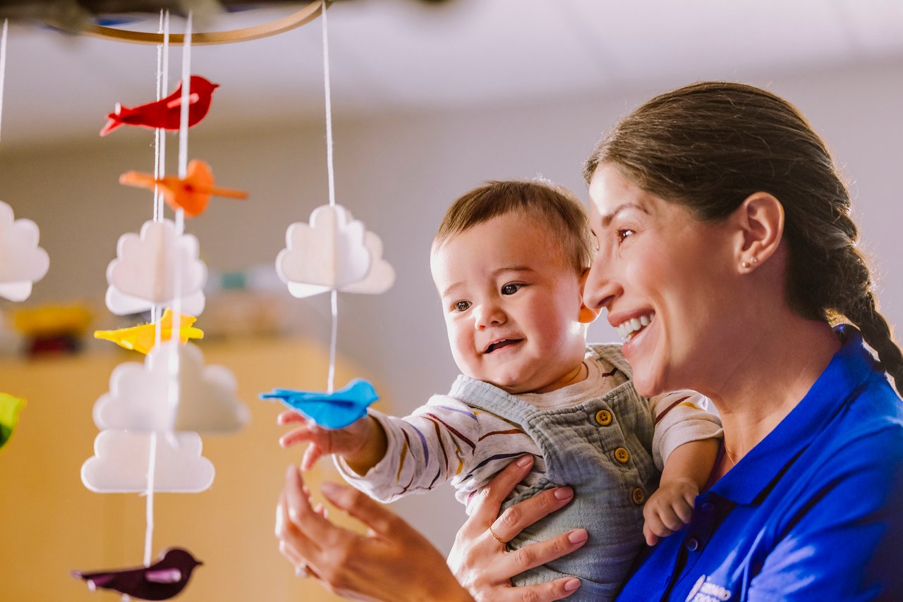 A teacher holding a baby up to a bird and cloud mobile in a preschool classroom
