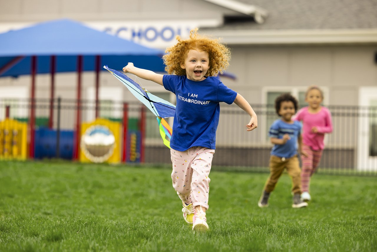 A child with curly red hair smiling and running with a kite across a preschool playground