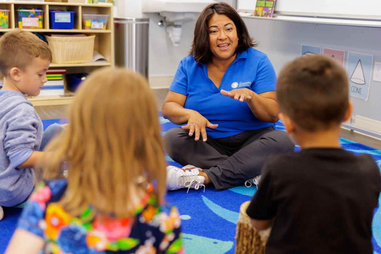 A teacher sitting on the floor talking to young children in a preschool classroom