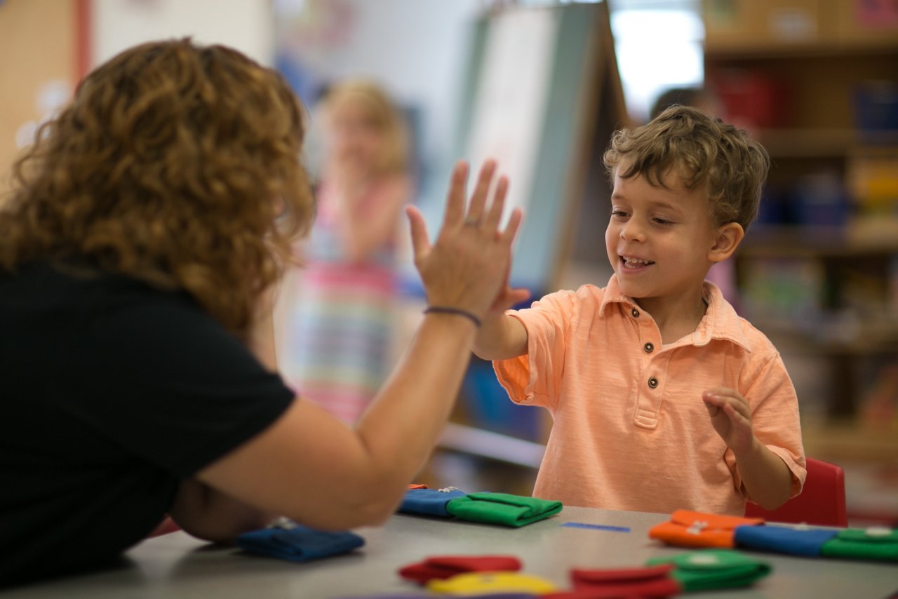 A young boy sits at a table and high-fives a teacher.