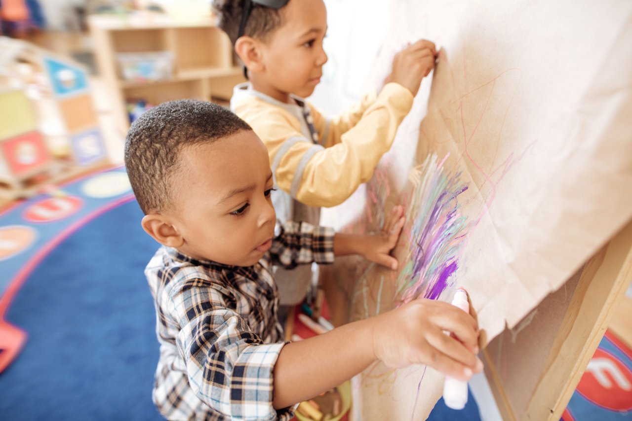 Two children standing and drawing with colorful markers on a large vertical piece of paper