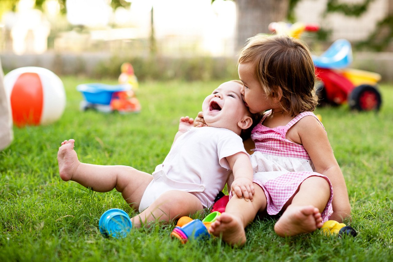 Toddler girl kissing baby sister on head while playing in the grass outdoors