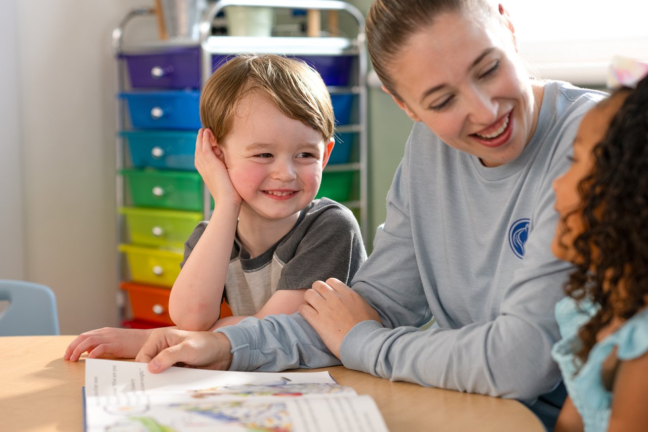 A joyful scene with a smiling teacher and two children at a table in a preschool classroom. Bright, colorful drawers are in the background, creating a warm, cheerful atmosphere.
