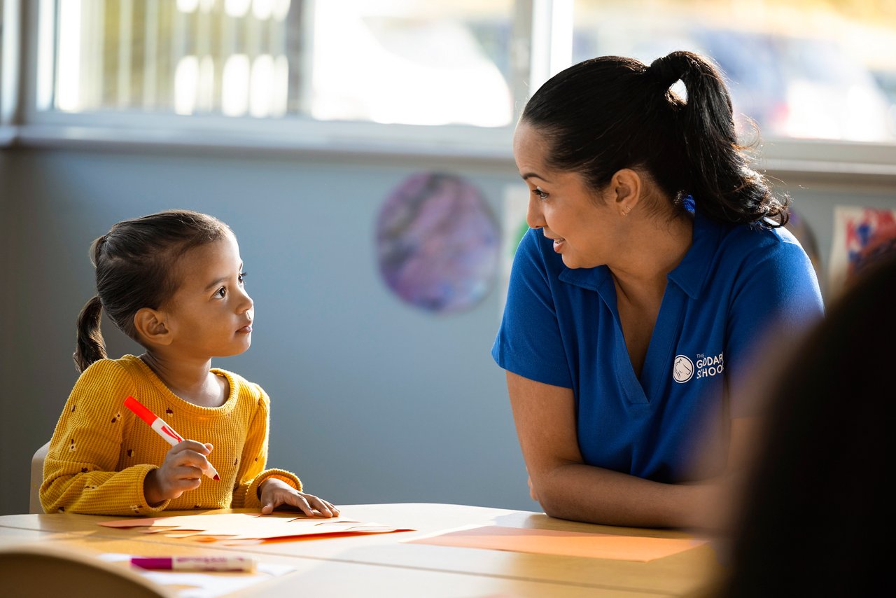A teacher speaking with a young girl who is coloring with a marker at a table in a preschool classroom.