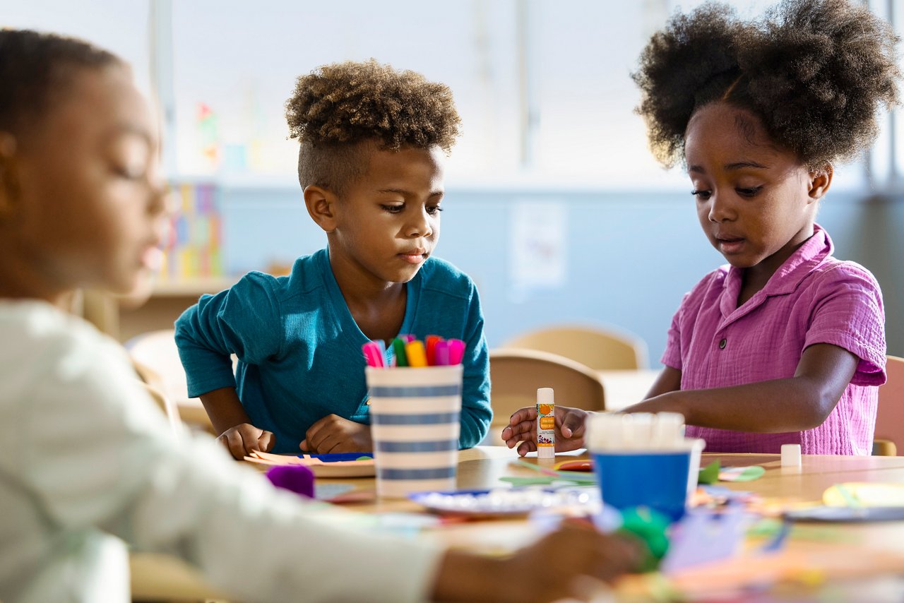 Young children crafting with markers, paper and glue sticks at a table in a preschool classroom.