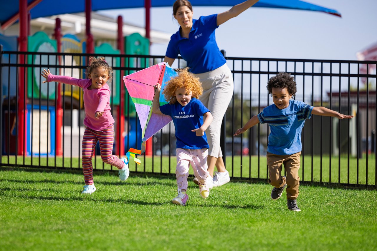 Three young children and a teacher running with a kite on a Goddard School playground.