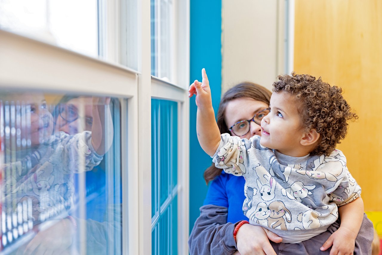 Caregiver holding a curious toddler pointing at a window, highlighting nurturing preschool environment and early childhood exploration.