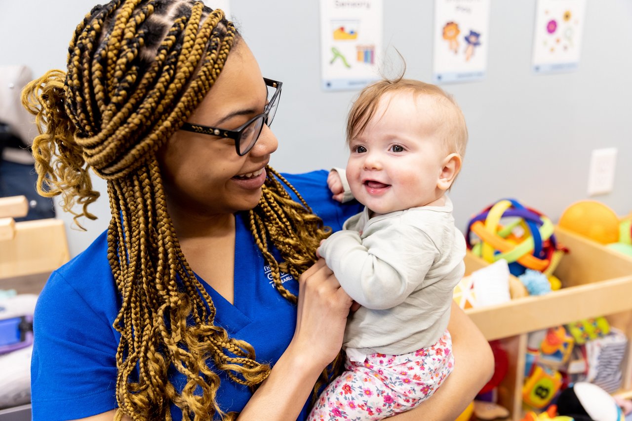 Caregiver holding an infant in a nurturing preschool classroom with toys and educational posters, emphasizing loving infant care and early learning environment.