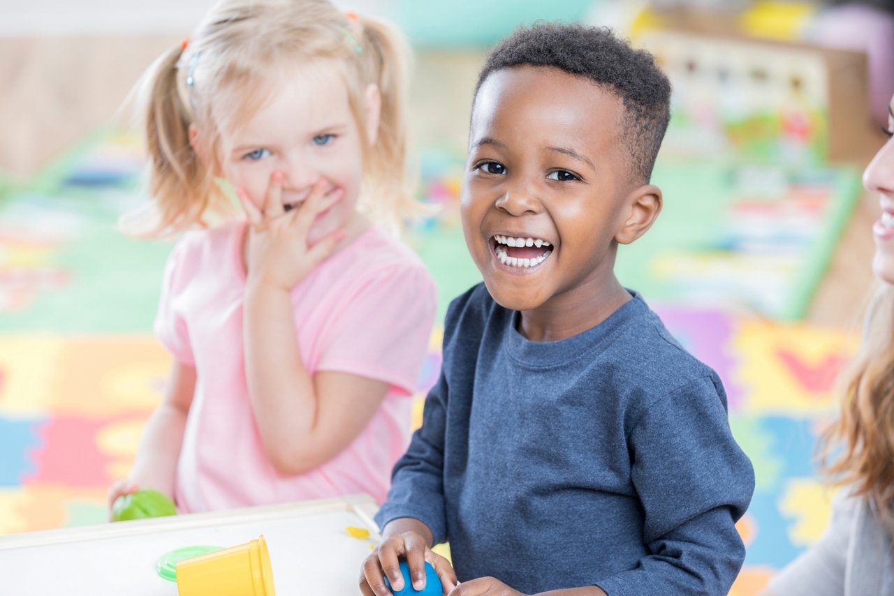 Adorable African American preschool boy smiles cheerfully as he plays with his friend in his preschool classroom.