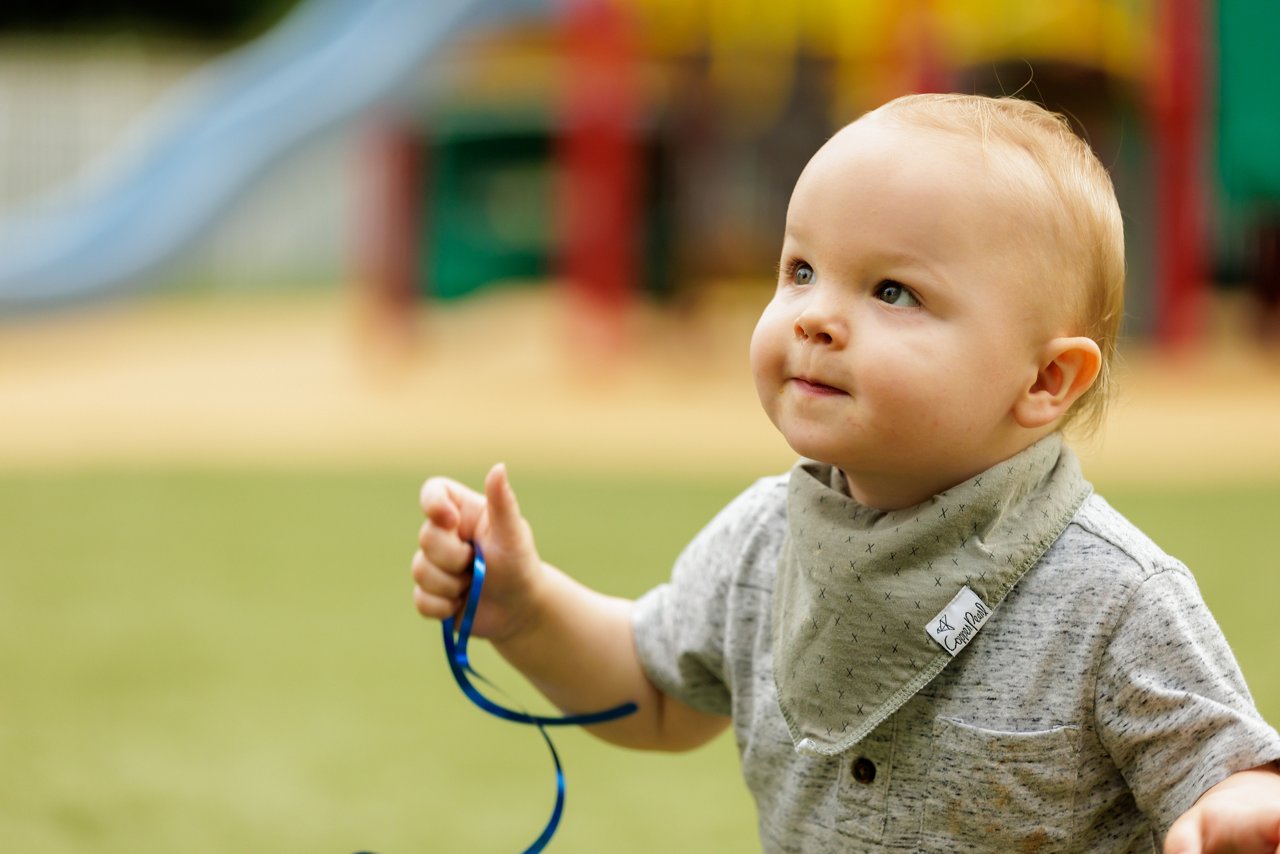Preschool child exploring a colorful outdoor playground while holding a blue ribbon—encouraging sensory play, movement, and discovery in a safe early childhood environment.