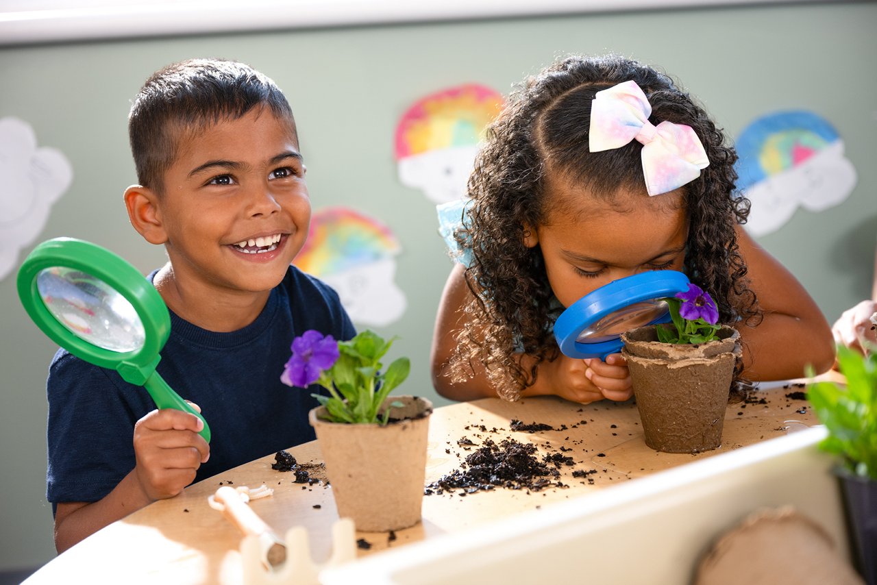 Two children are engaged with plants at a table. The boy smiles while holding a green magnifying glass, and the girl inspects a flower with a blue magnifying glass. Colorful drawings decorate the background, conveying a joyful and educational atmosphere.