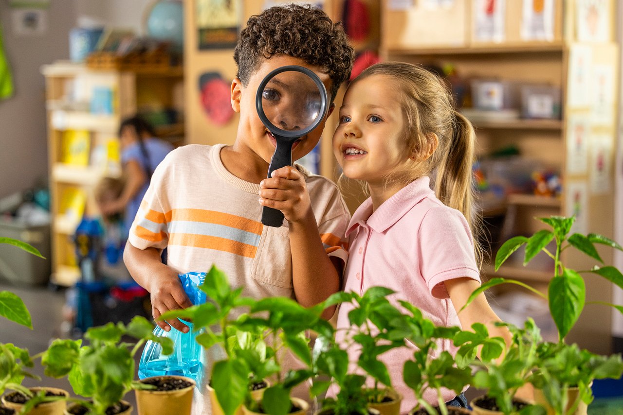 Two children in a preschool classroom explore potted plants. One holds a magnifying glass, and the other smiles. Bright, curious, and educational atmosphere.