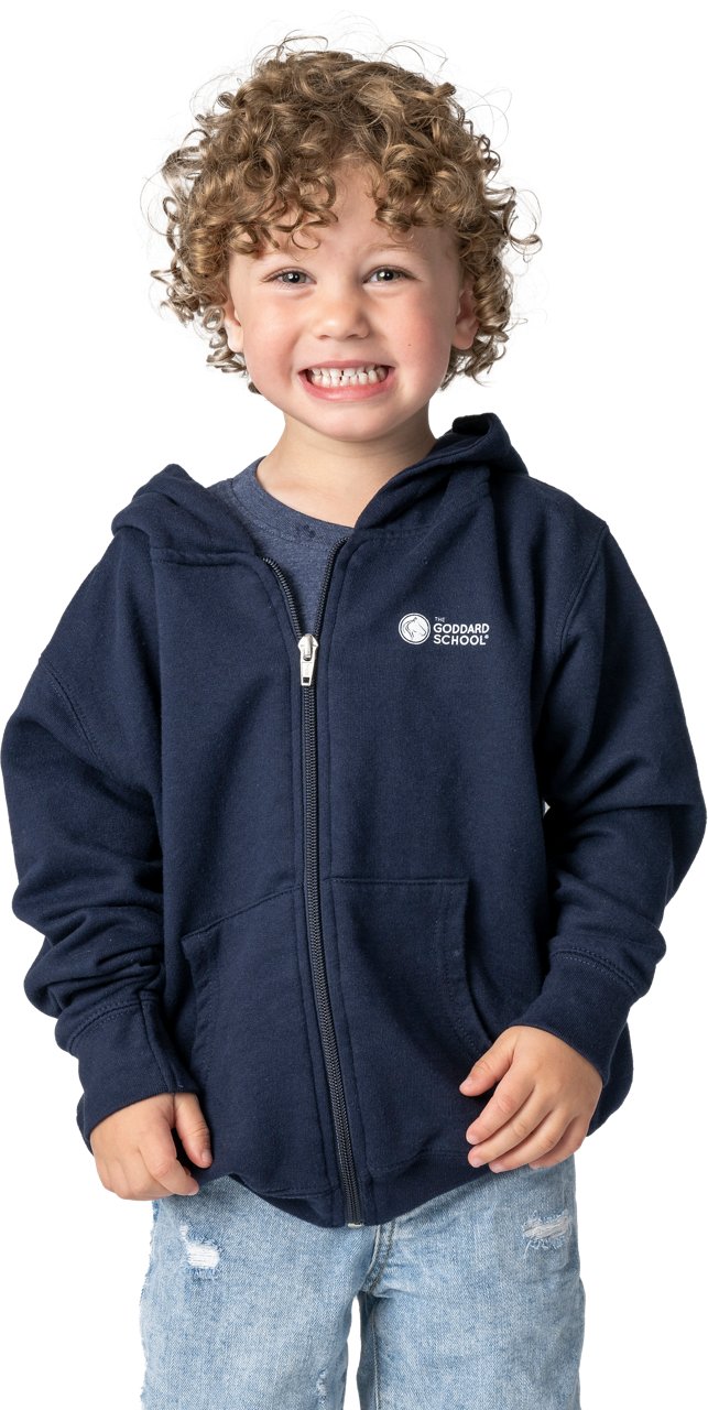 A smiling, curly-haired little boy wearing a navy blue Goddard School zip-up hoodie and jeans.