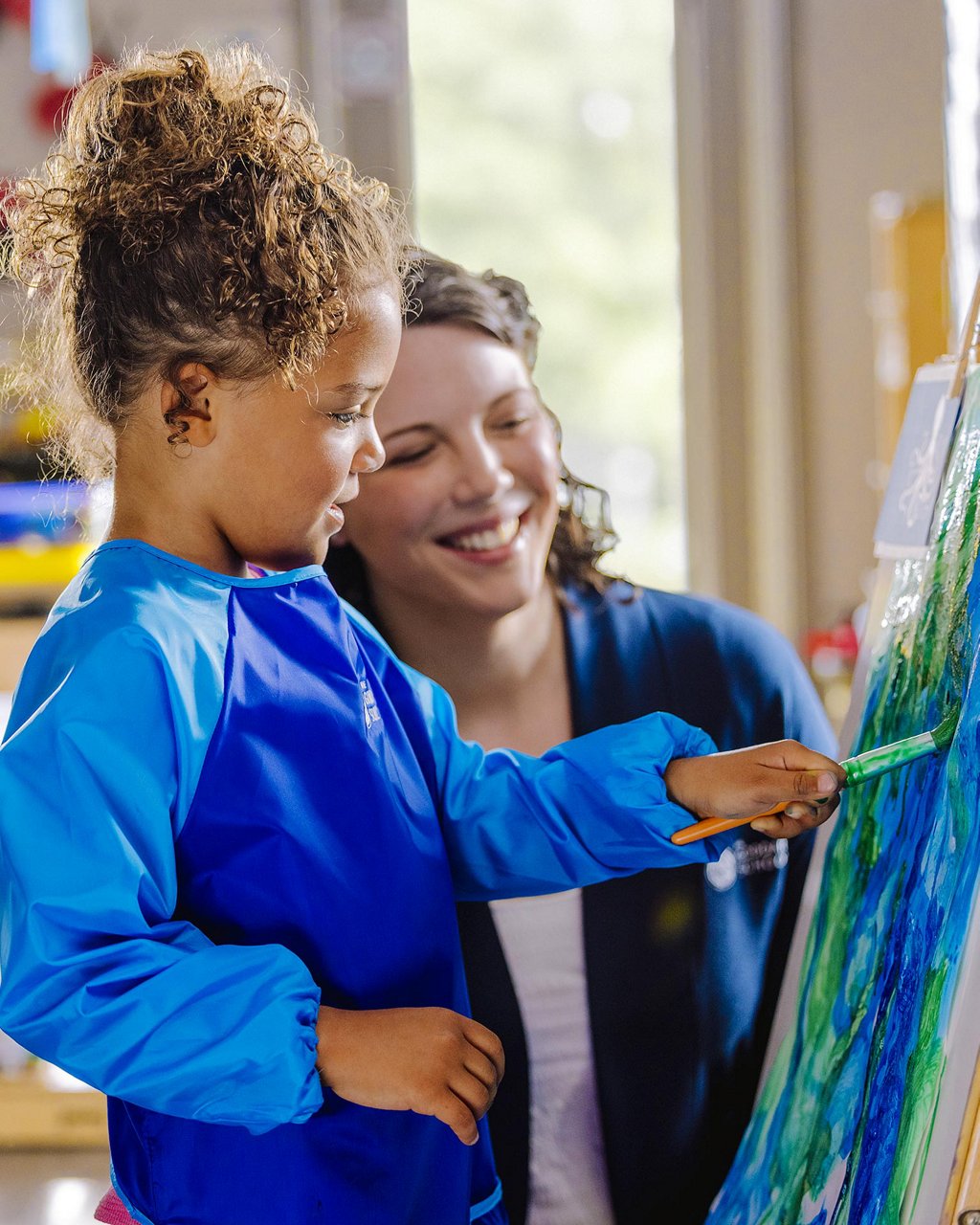 Young girl painting while teacher looks on proud and smiling