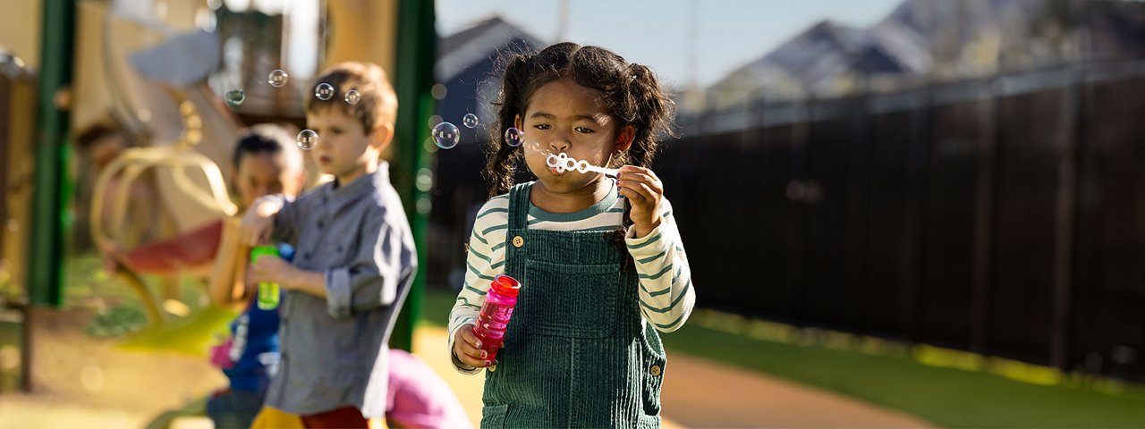 Young girl in overalls and other children blowing bubbles on a preschool playground