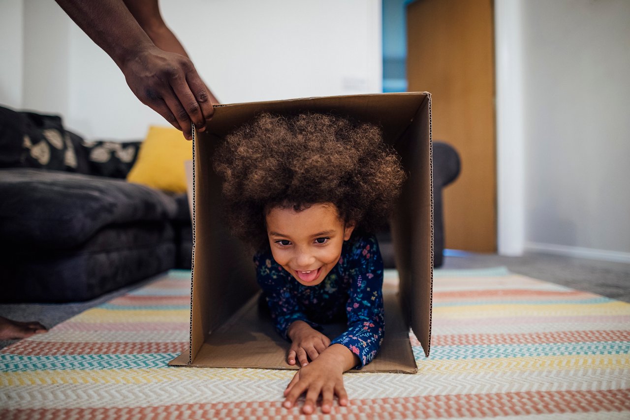 Mixed race girl crawling through a cardboard box in the sitting room at home, her unrecognisable father is holding the box up for her.