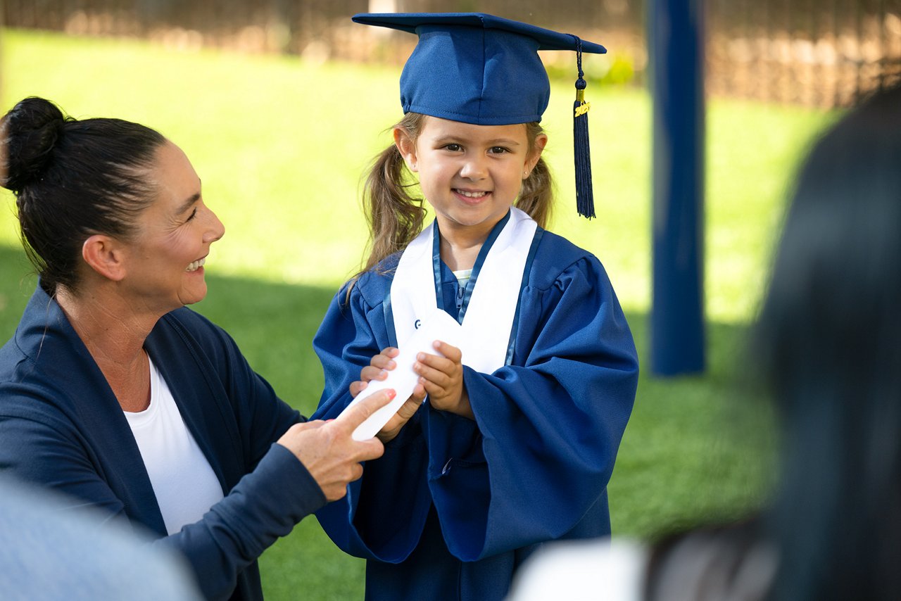 Young girl in blue graduation gown and cap, smiling, receiving a diploma from a woman. The scene is outdoors, conveying pride and joy.