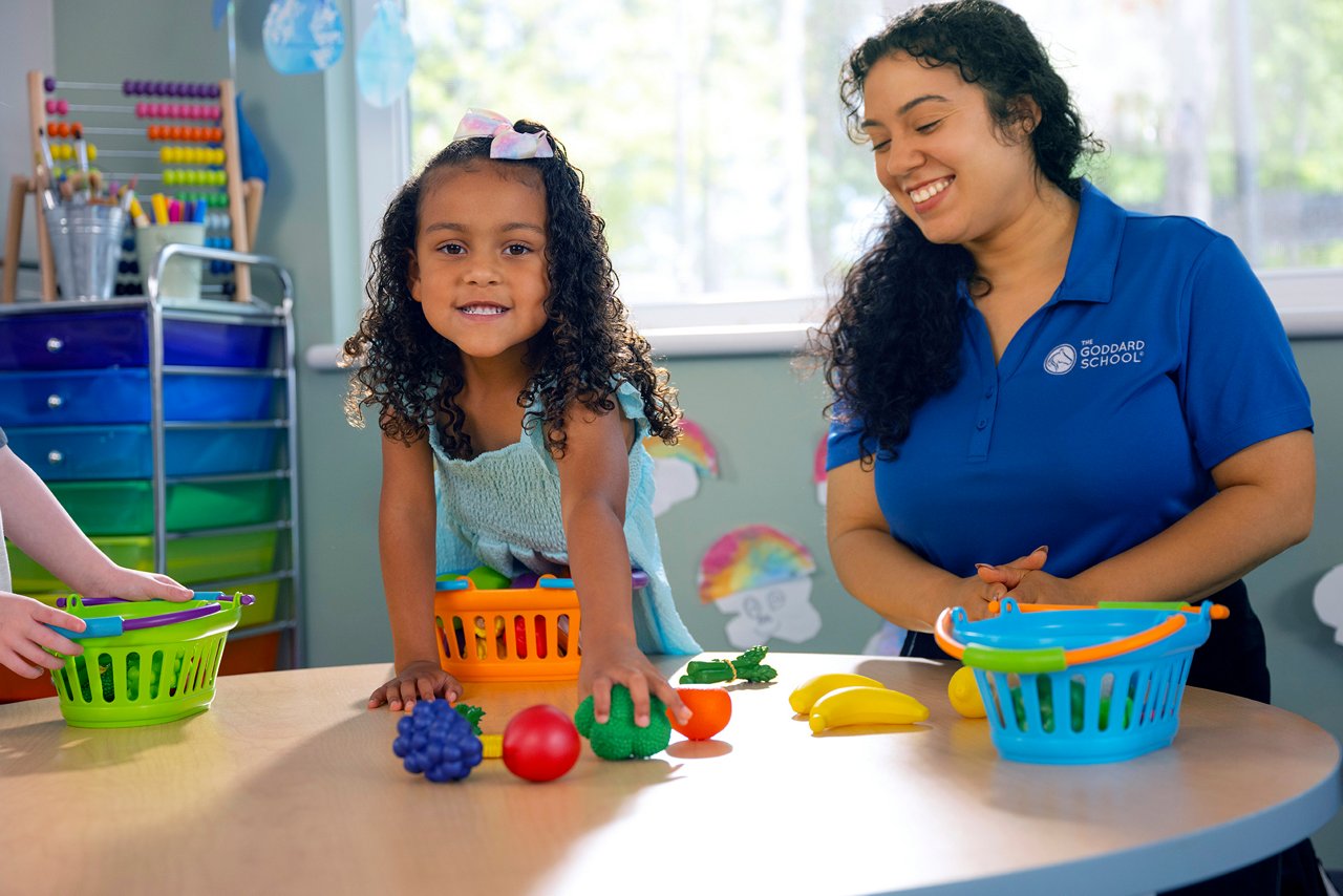 A cheerful child sorts colorful toy fruits into baskets at a table, assisted by a smiling teacher in a blue shirt. The preschool classroom is vibrant and inviting.