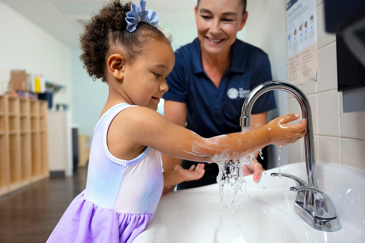 Young child washing hands with adult assistance under a faucet, promoting hygiene and handwashing habits in early childhood settings.