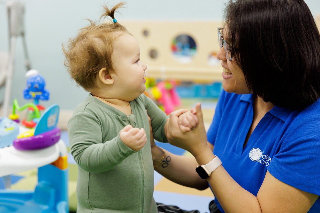 Infant standing with support from caregiver in a preschool play area, promoting safe infant care and early developmental milestones.