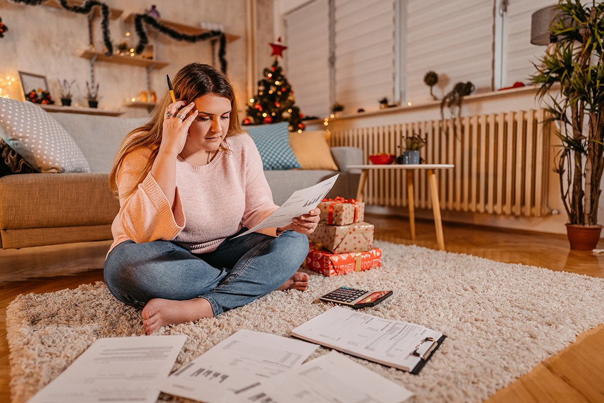 Person sitting on a rug reviewing holiday bills and budget papers with a calculator nearby, Christmas tree and wrapped gifts in the background.